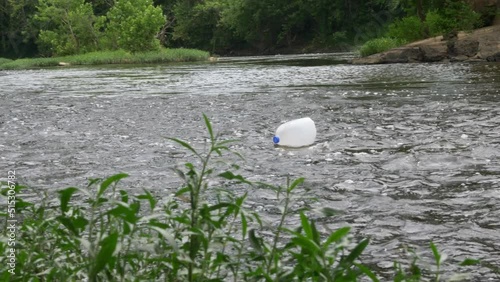 milk jug floats down river slowly behind foliage towards old concrete bridge on a cloudy day slow motion photo