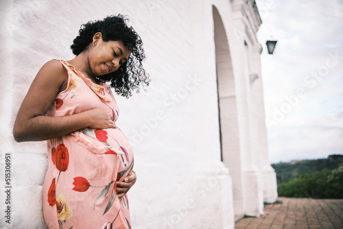 young african american pregnant latina in floral dress with white background looking at her belly