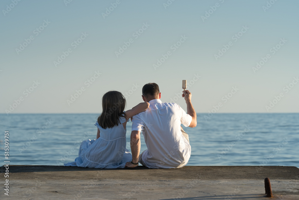 a girl and her dad are sitting on the pier and eating ice cream on a summer evening a girl and her dad are sitting on the pier and eating ice cream on a summer evening