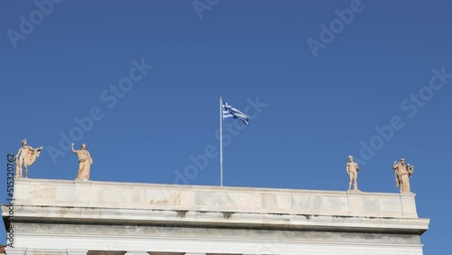 A roof of an old greek style building with two statues on each side of a flag pole with a waving greek flag on top. Ideal title scene for Greece travel video. photo