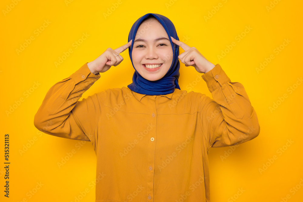 Smiling young Asian Muslim woman dressed in orange pointing to forehead, having great ideas isolated on yellow background Smiling young Asian Muslim woman dressed in orange pointing to forehead, having great ideas isolated on yellow background