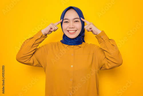Smiling young Asian Muslim woman dressed in orange pointing to forehead, having great ideas isolated on yellow background © Sewupari Studio