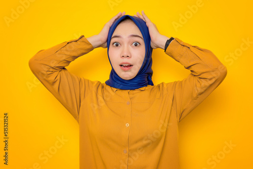 Shocked young Asian Muslim woman dressed in orange shirt looking at camera with open mouth and grabbing head isolated on yellow background © Sewupari Studio