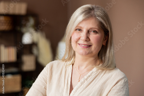 Headshot portrait positive mature woman with healthy smile standing on background of cozy living room, happy middle-aged lady looking to camera, posing for photo at home, enjoying leisure time.