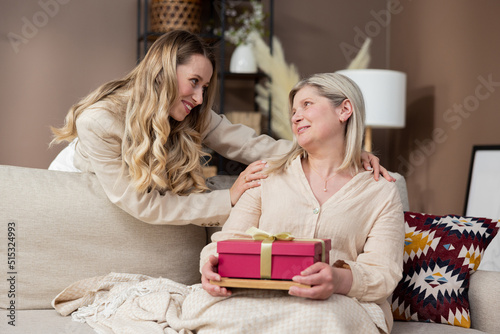 A beautiful adult woman hands a gift box to her mature mother and smiles as she sits on a couch in an elegant boho living room at home.