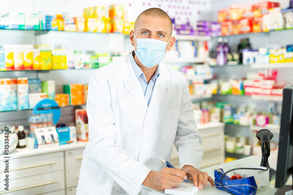 Caucasian male pharmacist in lab coat and face mask standing at counter in chemists shop and writing recipe.