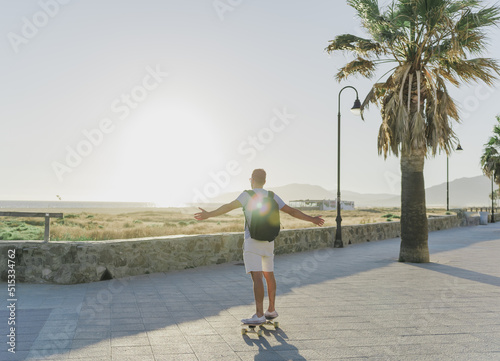 photo of an unrecognizable young man with his arms outstretched as he skates along the promenade at sunset