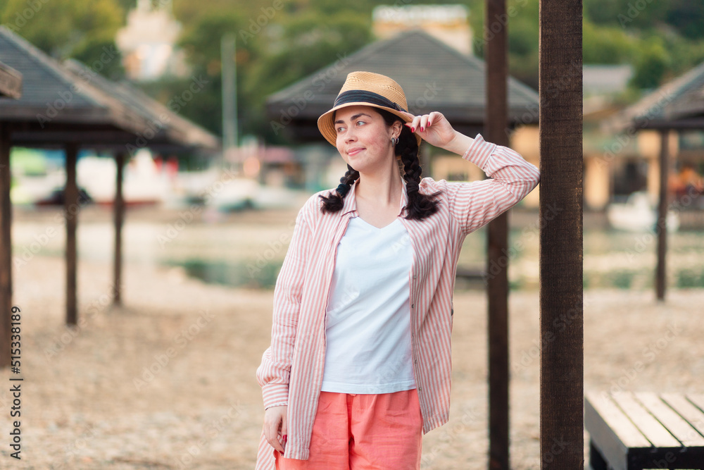 A young pretty Caucasian woman in summer clothes and a straw hat posing near beach umbrella. The concept of summer vacation