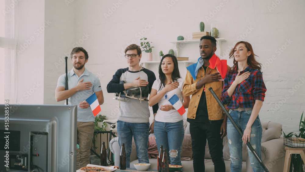 © silverkblack - Multi ethnic group of friends sport fans listening and singing French national anthem before watching sports championship on TV together at home indoors
