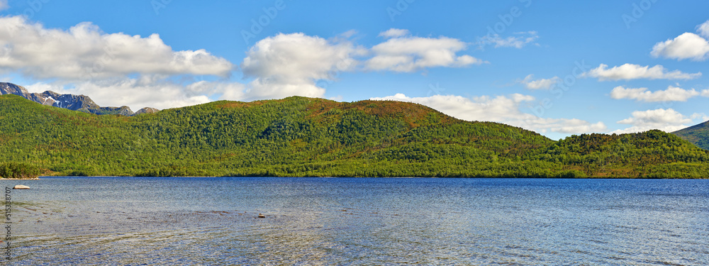 A hill behind a lake on summer day with clouds in the sky. Beautiful landscape view with greenery in nature. Mountain pastures in the background of a lake. Lush hill with pasture near water outdoors