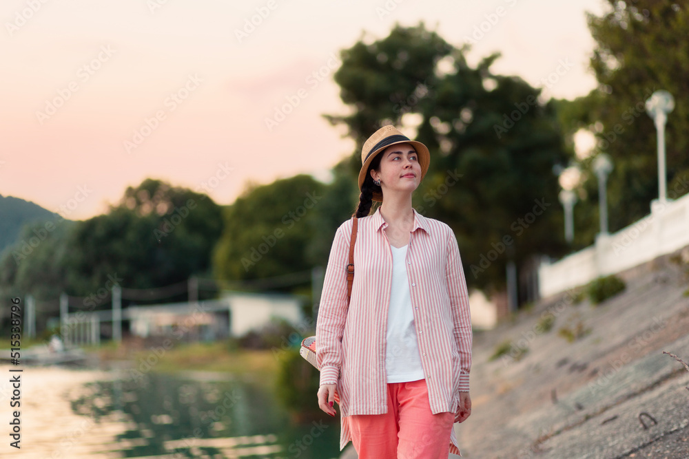 Summer vacation. Portrait of young attractive woman in summer clothes and straw hat walking down at river embankment. Evening stroll