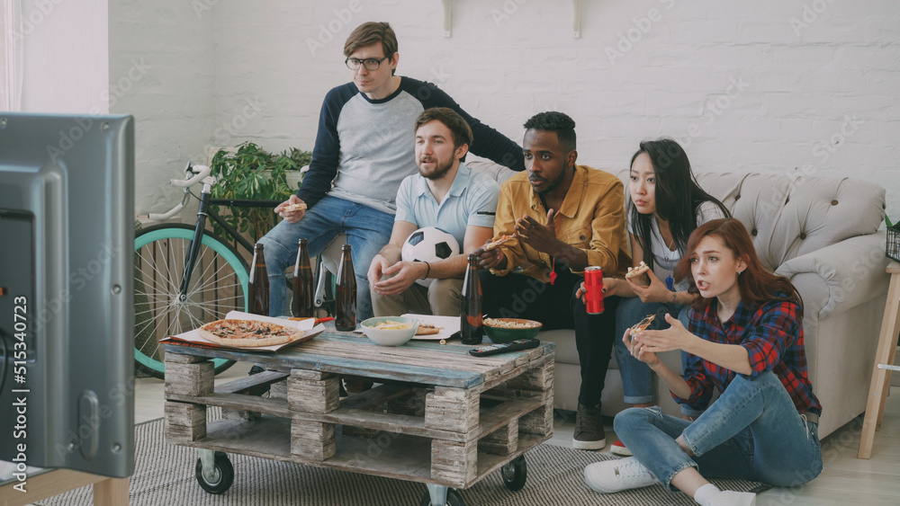 Multi-ethnic group of friends sports fans watching football championship on TV together eating pizza and drinking beer at home indoors Multi-ethnic group of friends sports fans watching football championship on TV together eating pizza and drinking beer at home indoors