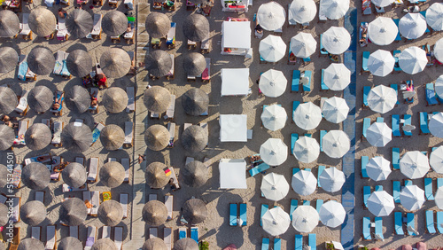Pattern made of densely arranged cane and white canvas umbrellas on the beach with people resting. Aerial shot. Summer background.