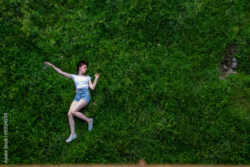 Portrait of happy cute young woman with short hair in casual black and white striped shirt, red round eyeglasses lying down on green grass and looking at camera through glasses.outdoor summertime shot