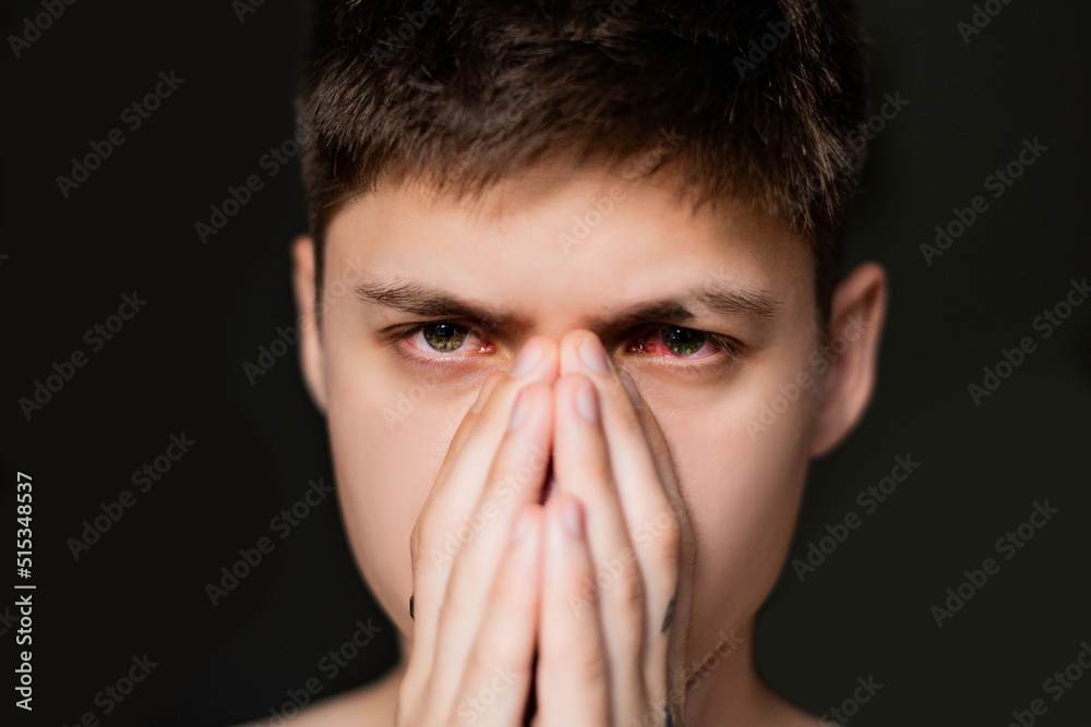 Young man crying on a black background. Boy closes his eyes with his hands. Close up of two annoyed red blood eyes of male affected by conjunctivitis or after flu, cold or allergy. Young man crying on a black background. Boy closes his eyes with his hands. Close up of two annoyed red blood eyes of male affected by conjunctivitis or after flu, cold or allergy.