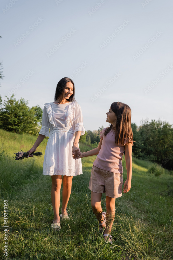 Fototapeta premium happy mother and daughter holding hands while walking on rustic road on summer day.