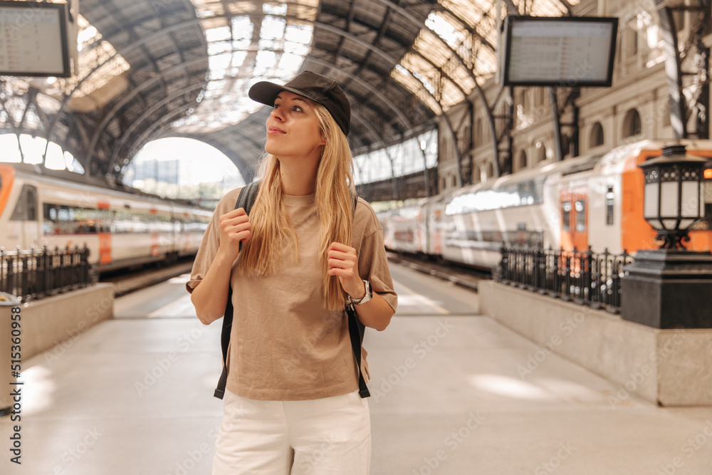 Fototapeta premium View of caucasian fair-skinned woman at the train station . Focused on blonde looking away in black cap with backpack 