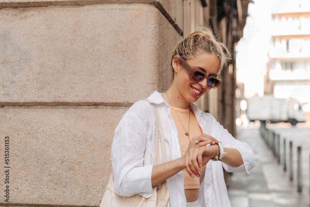 Caucasian blonde woman holding her watches on hand and checking time. Smiling fair skinned in sunglasses at the street 