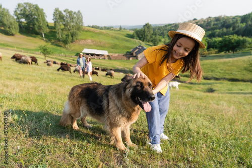 girl in straw hat cuddling cattle dog near parents herding livestock on blurred background. © LIGHTFIELD STUDIOS
