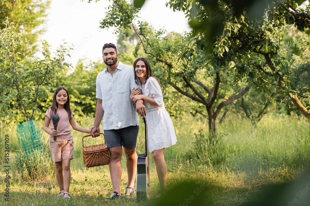 Fototapeta premium joyful family with wicker basket and guitar looking at camera in countryside garden.