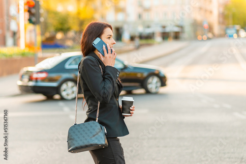 Young business woman talking on the phone holding coffee paper cup,crossing street blurred background. Fashion business photo of beautiful girl in black suite with phone and cup of coffee.Side view. © ARVD73