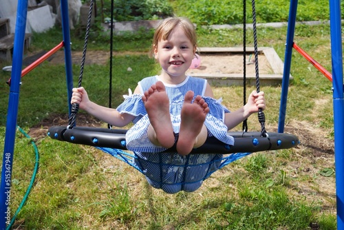 Happy little girl dressed in a blue striped dress is enjoying a swing ride on a sunny summer playground in the garden.