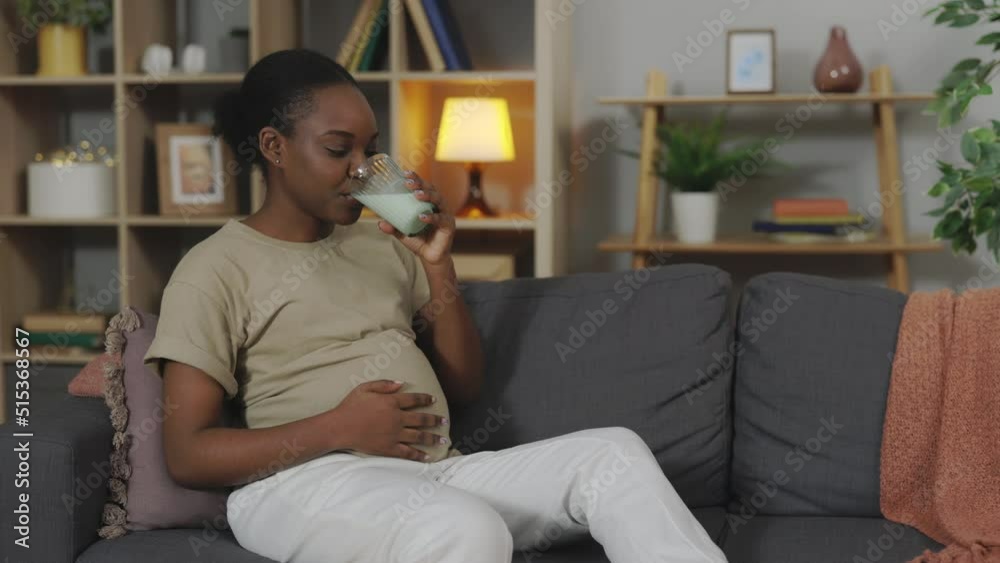 African american woman in expectation wearing casual clothes drinking tasty milk from glass while sitting on sofa. Concept of healthy eating during pregnancy. African american woman in expectation wearing casual clothes drinking tasty milk from glass while sitting on sofa. Concept of healthy eating during pregnancy.