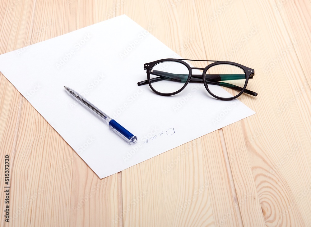 Exquisite black glasses are arranged on a light wood structural craft and texture office table next to a sheet of white paper with a pen on the smooth surface of the paper. No people photography. Exquisite black glasses are arranged on a light wood structural craft and texture office table next to a sheet of white paper with a pen on the smooth surface of the paper. No people photography.