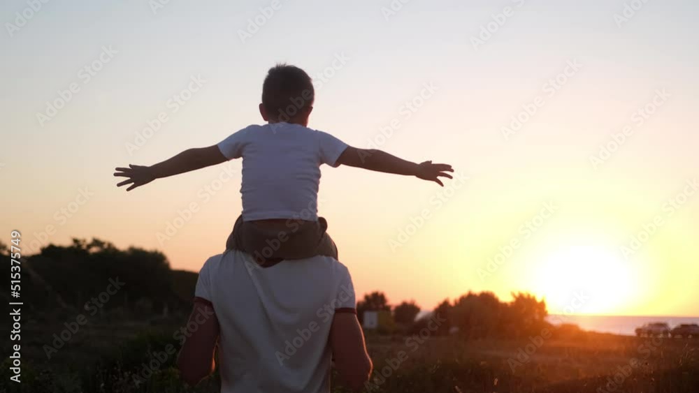 Dad carries a little son on his shoulders walking across the field against the backdrop of a sunset in the sky, back view, slow motion Dad carries a little son on his shoulders walking across the field against the backdrop of a sunset in the sky, back view, slow motion