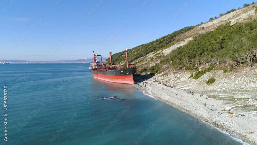 Naklejka premium Aerial for an empty industrial ship moored near sea shore with many people walking on a beach. Maritime cargo vessel standing near green trees slope in a summer sunny day.