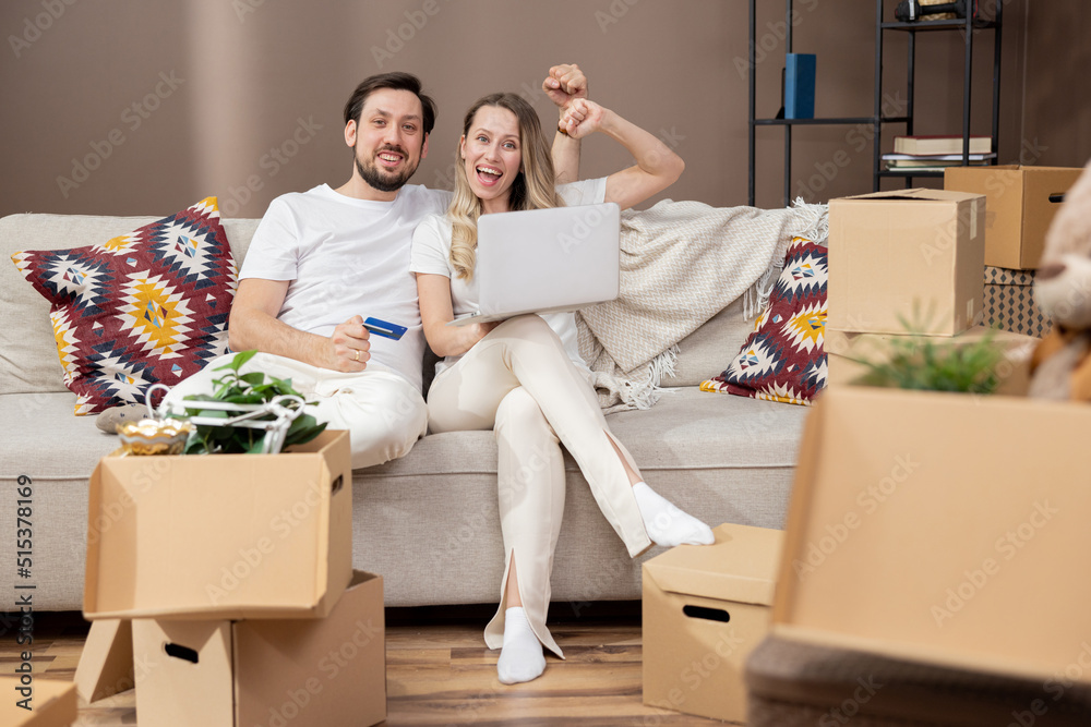 Portrait of a laughing young couple shopping online, sitting on a couch with a credit card and laptop in front of them, a stack of cardboard boxes from the move. Portrait of a laughing young couple shopping online, sitting on a couch with a credit card and laptop in front of them, a stack of cardboard boxes from the move.