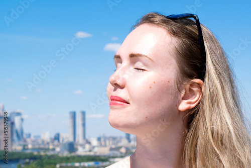 Young woman with blond hair against the blue sky and cityscape. The woman is enjoying the sun and fresh air. Concept of maintaining healthy and fresh skin in a big city