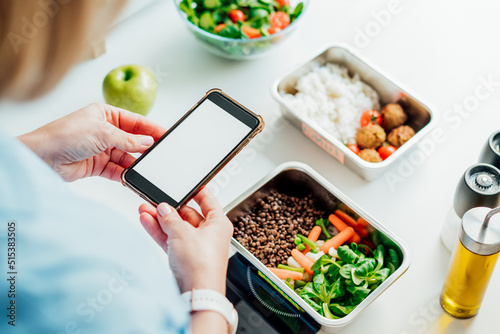 Healthy diet plan for weight loss, daily ready meal menu. Woman using phone with blank screen for app while weighing lunch box cooked in advance on kitchen scale. Balanced portion. Pre-cooking