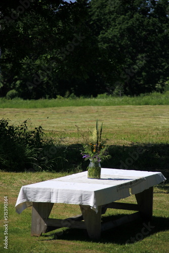 A large wooden table with a white tablecloth and a vase of meadow flowers stands in a green meadow under large trees on the shore of a lake, a Summer Solstice food table in the yellow sunset light