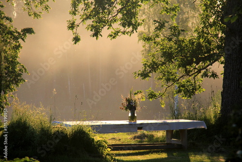 A large wooden table with a white tablecloth and a vase of meadow flowers stands in a green meadow under large trees on the shore of a lake, a Summer Solstice food table in the yellow sunset light
