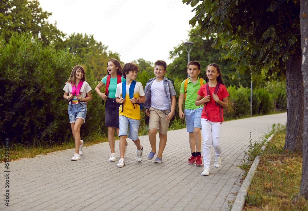 Back to school. Cheerful group of junior high school students with backpacks on their shoulders walk on sidewalk in park. Joyful children in summer casual clothes go and talk to each other on warm day