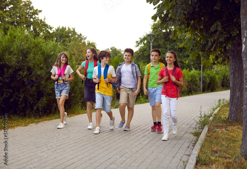 Back to school. Cheerful group of junior high school students with backpacks on their shoulders walk on sidewalk in park. Joyful children in summer casual clothes go and talk to each other on warm day