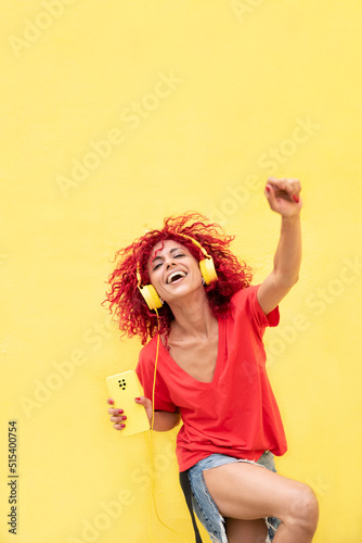 happy latin woman with afro red hair holding a smartphone,dancing and listening to music with yellow headphones over yellow background wearing red shirt