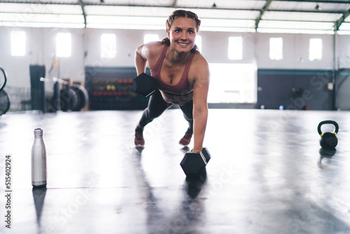 Happy female with dumbbells training strength doing plank exercise in gym, smiling woman working on hands and chest muscles pushing from floor in gym studio keeping perfect body shape and vitality