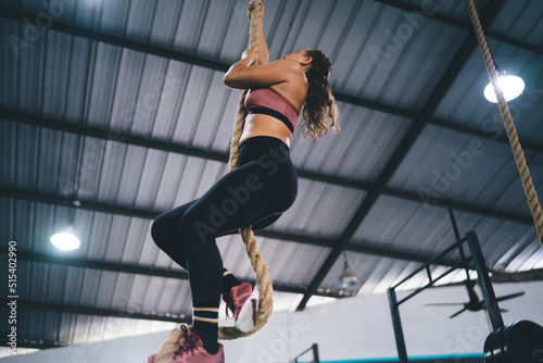 Pumped up female athlete in sportswear climbing at rope simulator with bodyweight during hard workout training in gym studio, Caucaisan woman in tracksuit slimming and keeping figure in good shape
