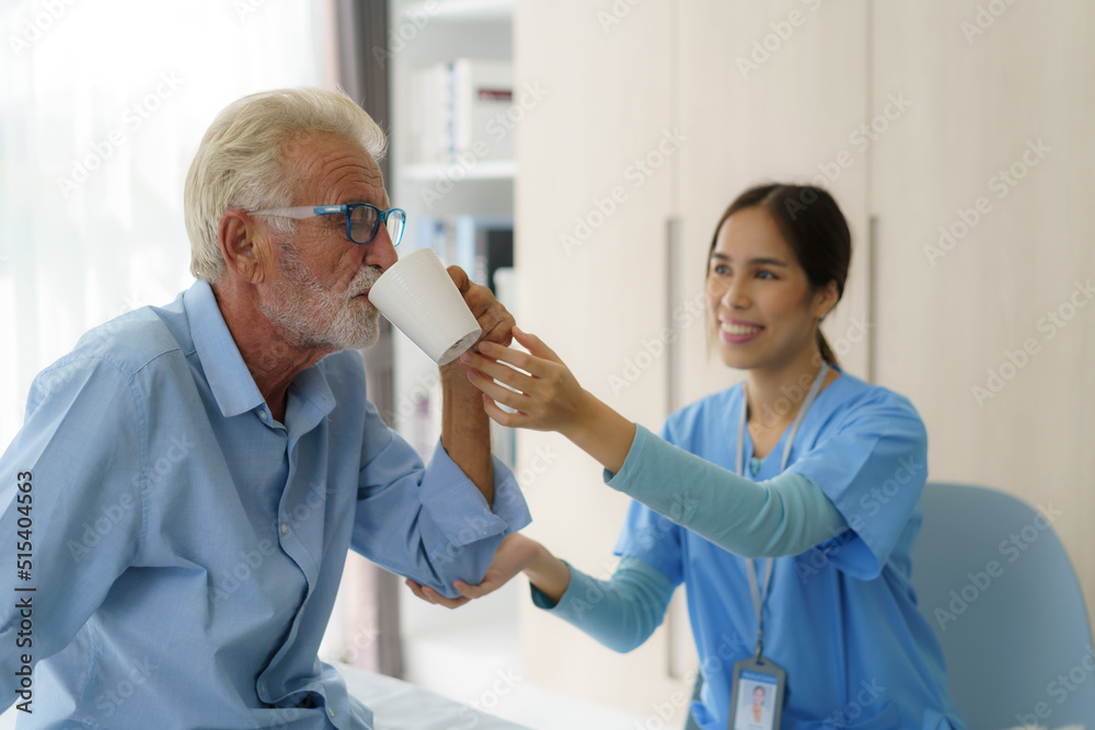 Fototapeta premium Asian elderly caregiver woman sitting on a hospital bed next to an older man helping drink a glass of water from the bedroom at home.