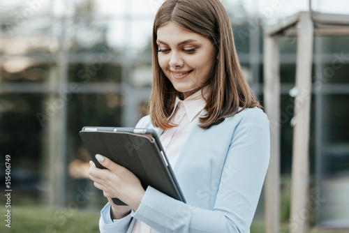 Businesswoman holding tablet in hands using business apps on tablet computer near office building. Elegant female chatting online with digital device outside. Happy face looking tablet screen outdoors