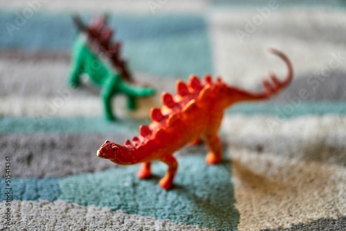 Fototapeta Naklejka Na Ścianę i Meble -  Closeup shot of a red stegosaurus toy on the carpet with a dinosaur toy in the blurred background