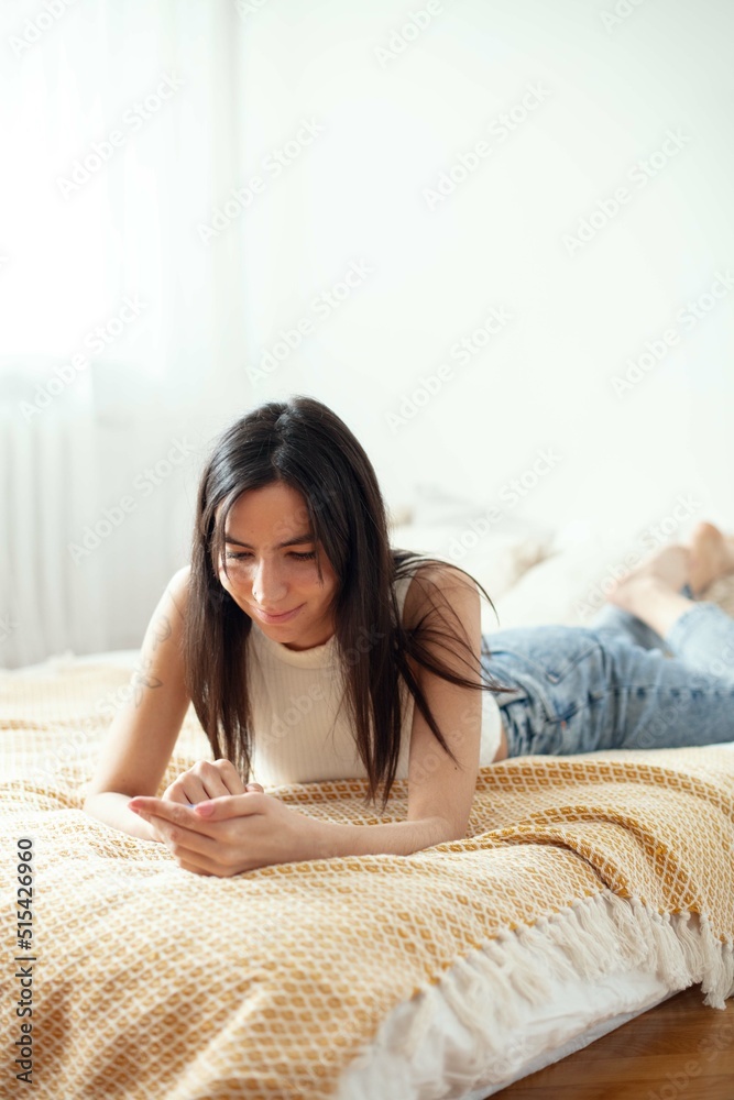Young Smiling Woman Using Phone While Lying On Sofa At Home.