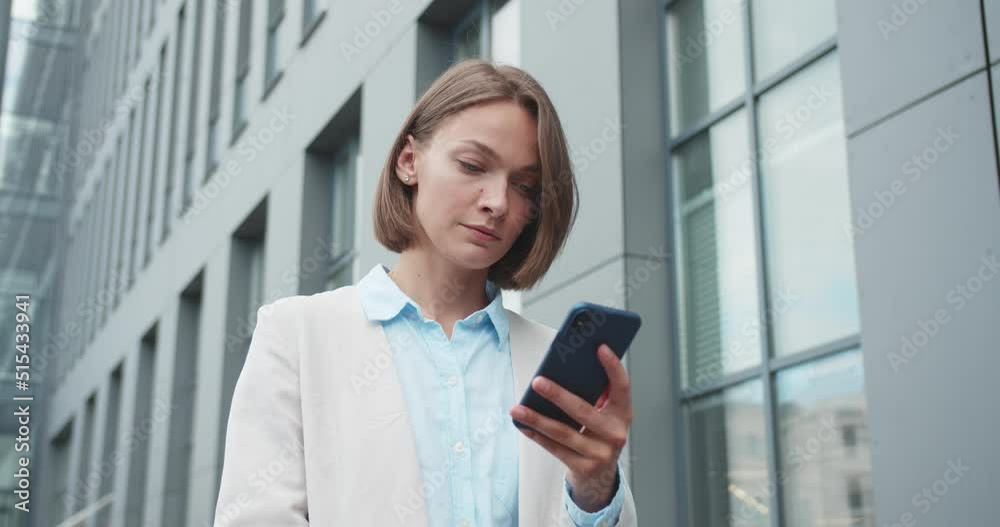 Beautiful young Caucasian woman standing in city street and texting on mobile phone. Businesswoman having telephone chat, messaging outdoors at office center and looking at camera. Portrait. Beautiful young Caucasian woman standing in city street and texting on mobile phone. Businesswoman having telephone chat, messaging outdoors at office center and looking at camera. Portrait.