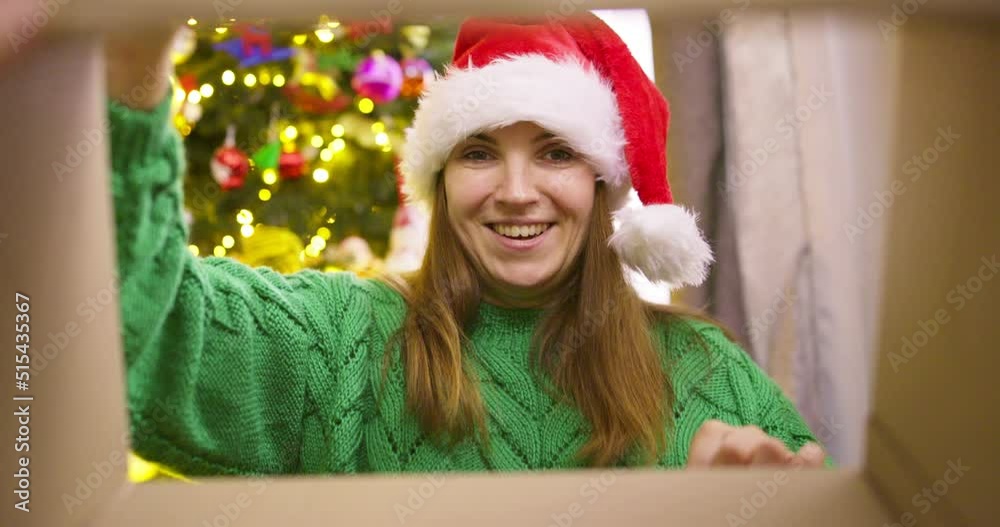 Beautiful young woman in a Christmas hat opening Christmas gift box with excited surprised face feeling happy on New Year's Eve sitting at home near glowing christmas tree. Magic moments Beautiful young woman in a Christmas hat opening Christmas gift box with excited surprised face feeling happy on New Year's Eve sitting at home near glowing christmas tree. Magic moments
