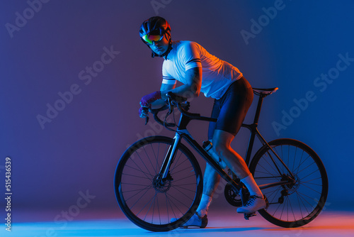 One male cyclist riding bicycle wearing cycling shorts and protective helmet isolated on dark blue background in neon. Concept of sport, speed, energy