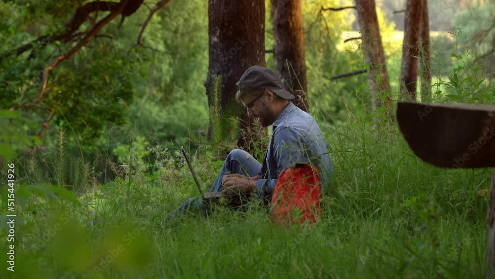 Happy Man tourist with backpack uses laptop outdoors in summer forest. Freelancer distant works while traveling in nature. Happy Man tourist with backpack uses laptop outdoors in summer forest. Freelancer distant works while traveling in nature.