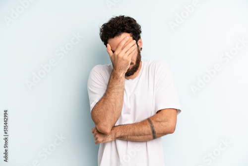 young adult bearded man looking stressed, ashamed or upset, with a headache, covering face with hand