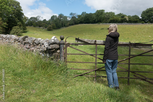 A lady walker looks across a farm field as she leans on an iron gate with a traditional Yorkshire dry stone wall in view
